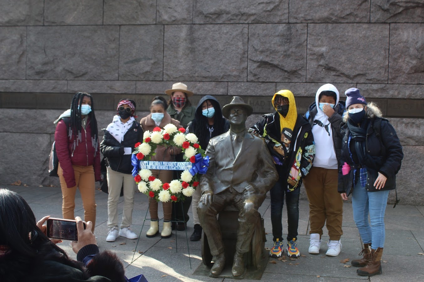 Students from Browne Education Campus surround FDR Wheelchair statue to take picture with Mary Dolan, executive director of FDR Committee, and the beautiful red, white, and blue wreath to the left of statue.
